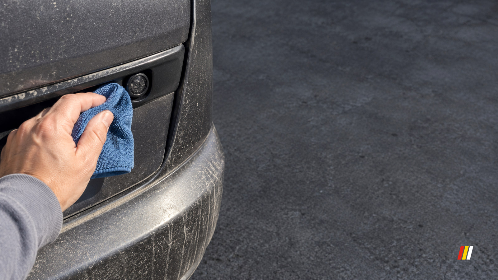 A hand holds a blue cloth against the dusty back of a car beside a small round camera, with gray pavement in the background.