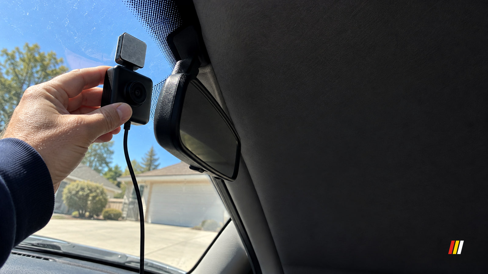 A hand holds a wired dash cam near the windshield and rearview mirror inside a car, with blue sky and a sunlit driveway visible outside.