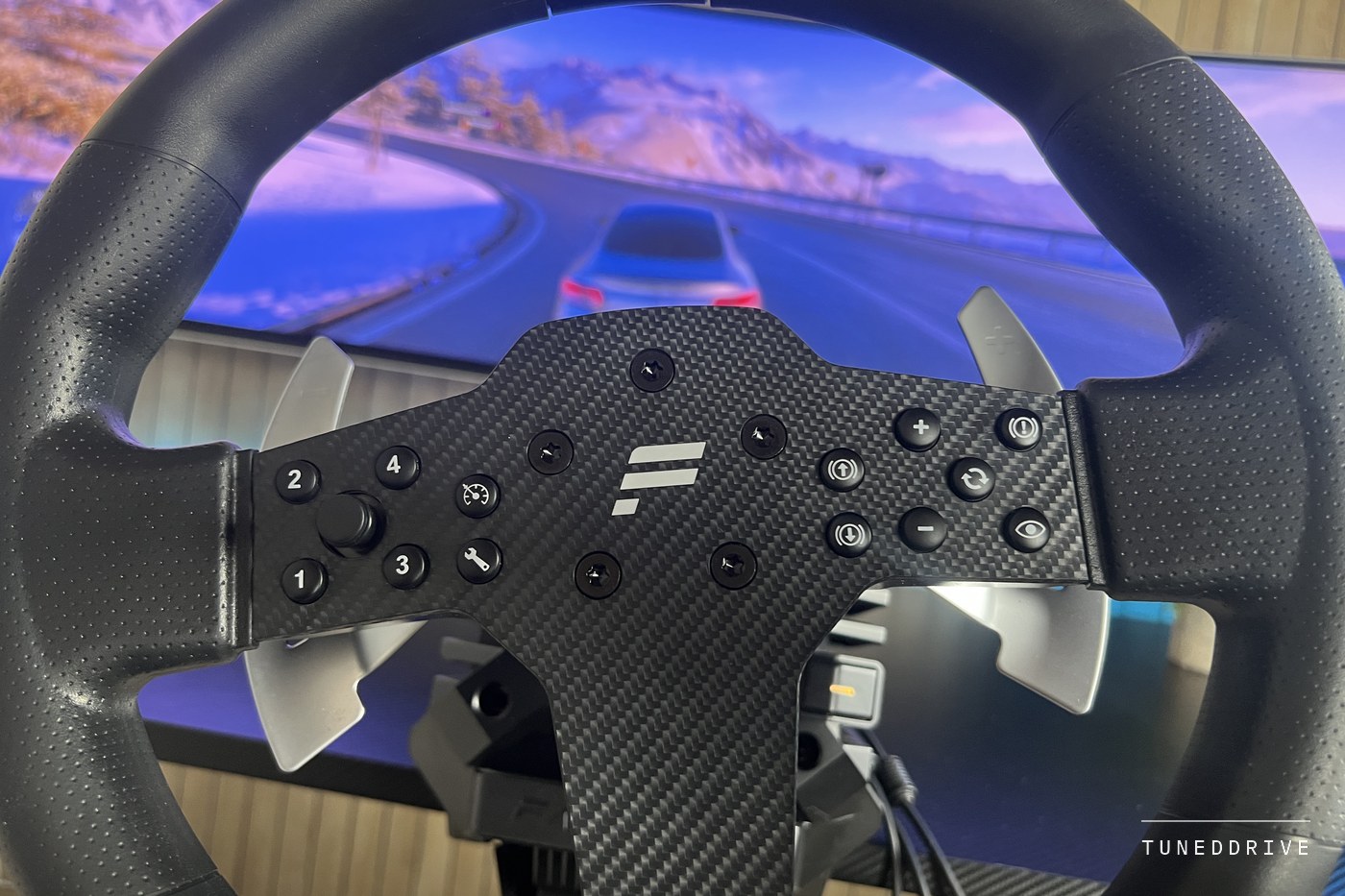 Close view of the P1 V2 wheel's centre with its buttons, directional stick and metal shift paddles, a racing game behind