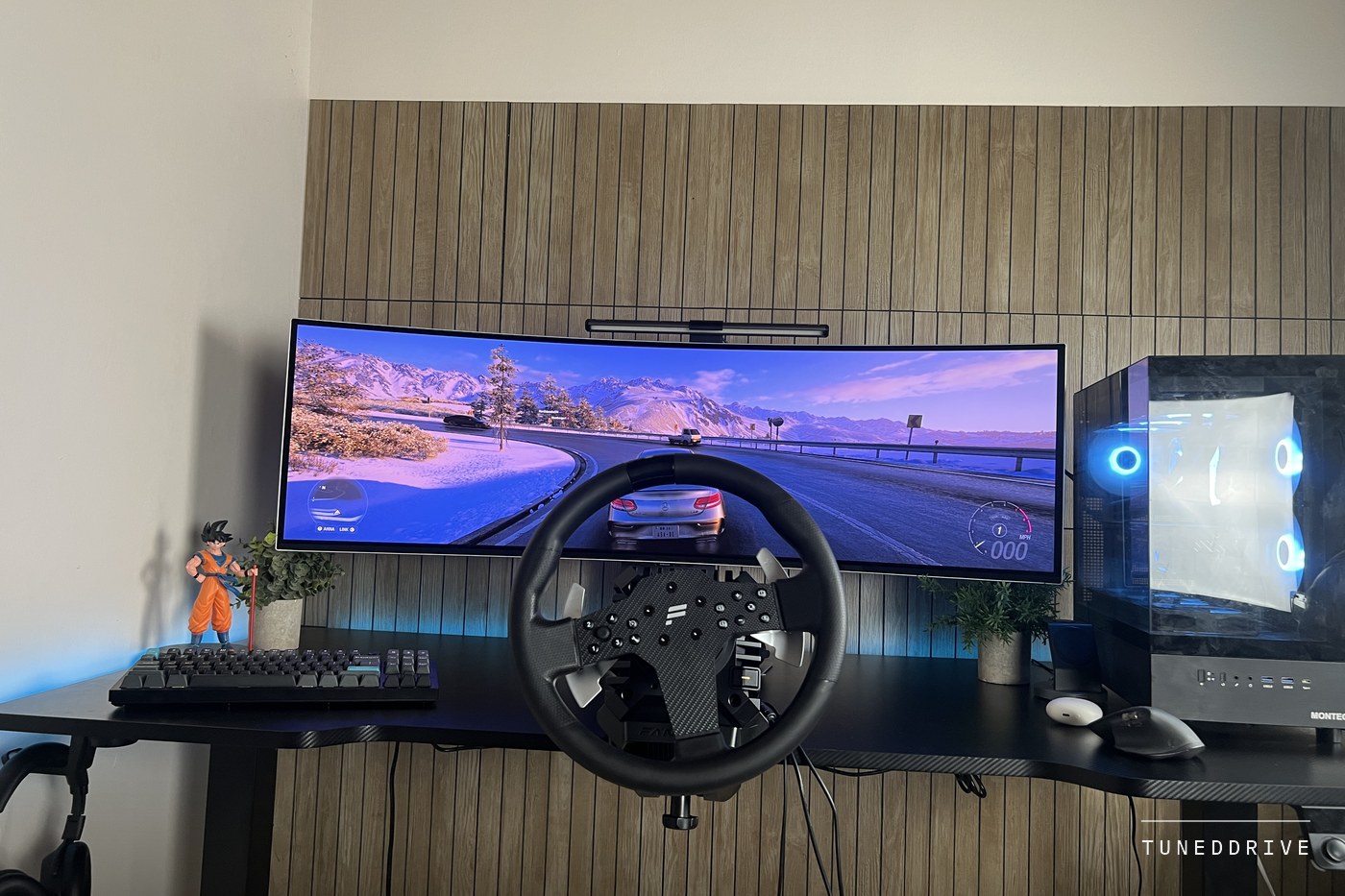 A desk setup with the Fanatec wheel and base in front of a curved ultrawide monitor, keyboard on the left and a PC tower on the right, slatted wood wall behind