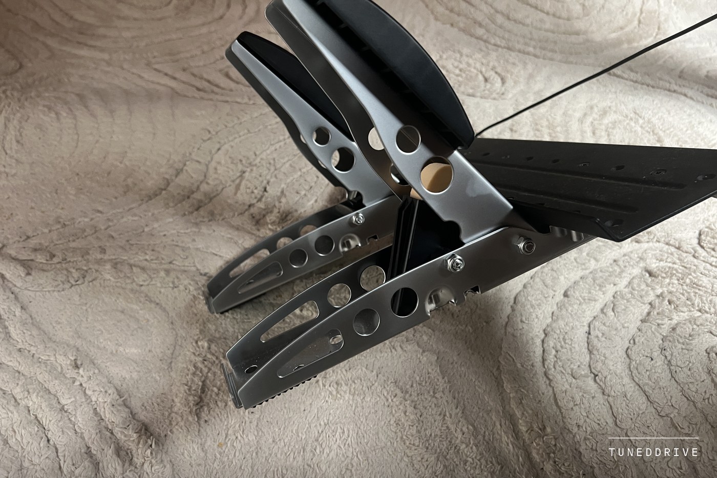 The Fanatec CSL throttle and brake pedals from the side on a cream rug, their steel arms cut with round holes