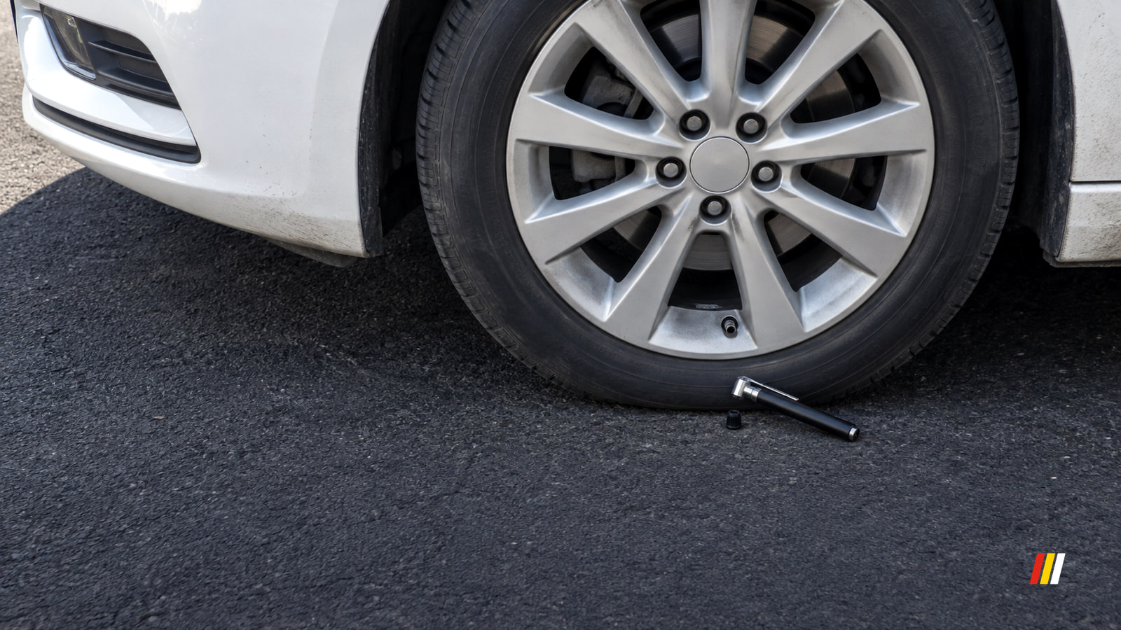 The front wheel of a white car in a car park, valve cap off, with a black tyre pressure gauge on the asphalt beside it in hard midday sun.