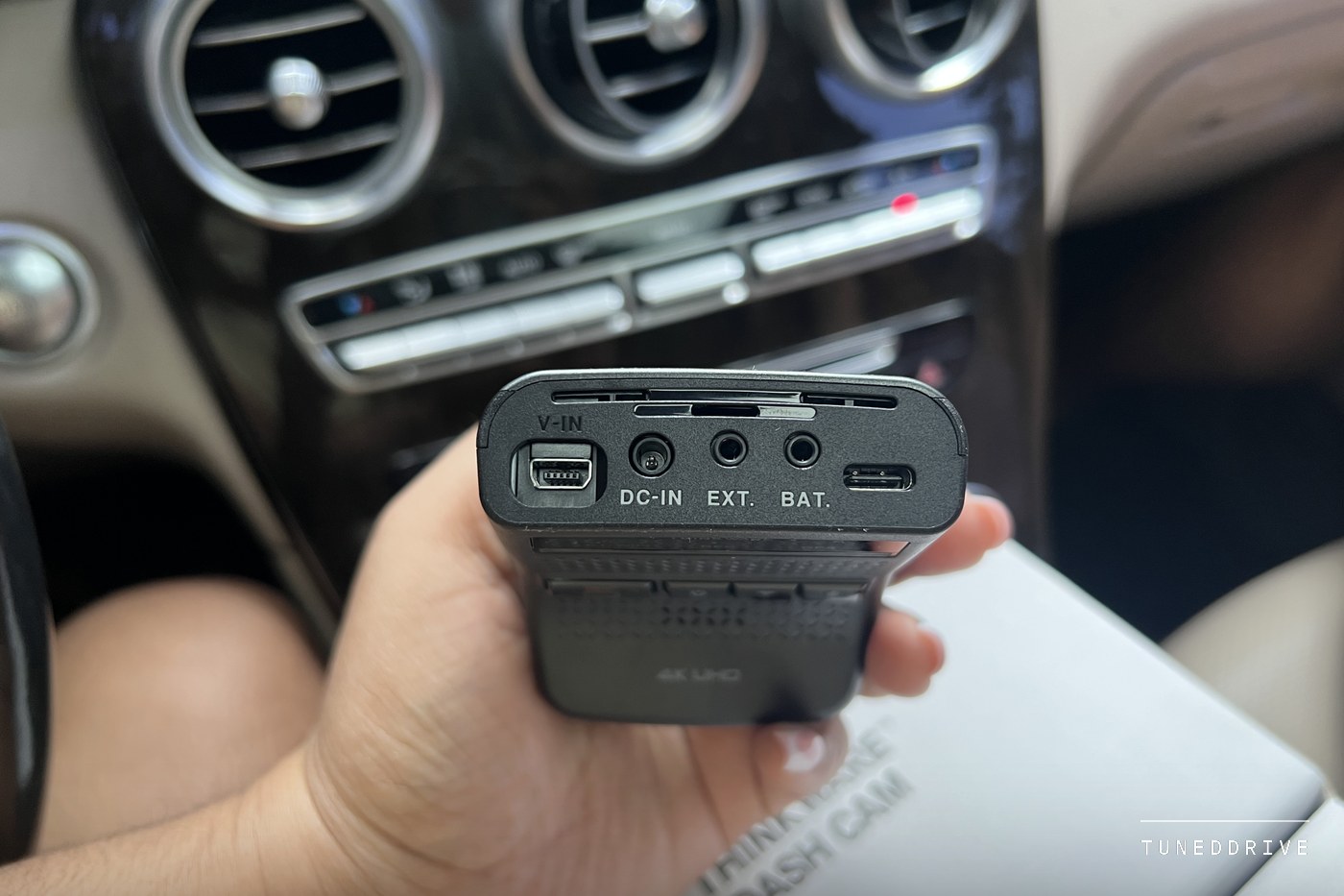 The edge of the camera body showing its ports labelled V-IN, DC-IN, EXT and BAT alongside a USB-C socket, held in front of the dashboard