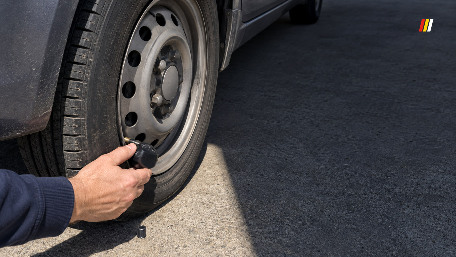 A hand holds a black pressure gauge on the valve of a dark grey car's slightly soft rear tire on a concrete driveway in hard midday sun.