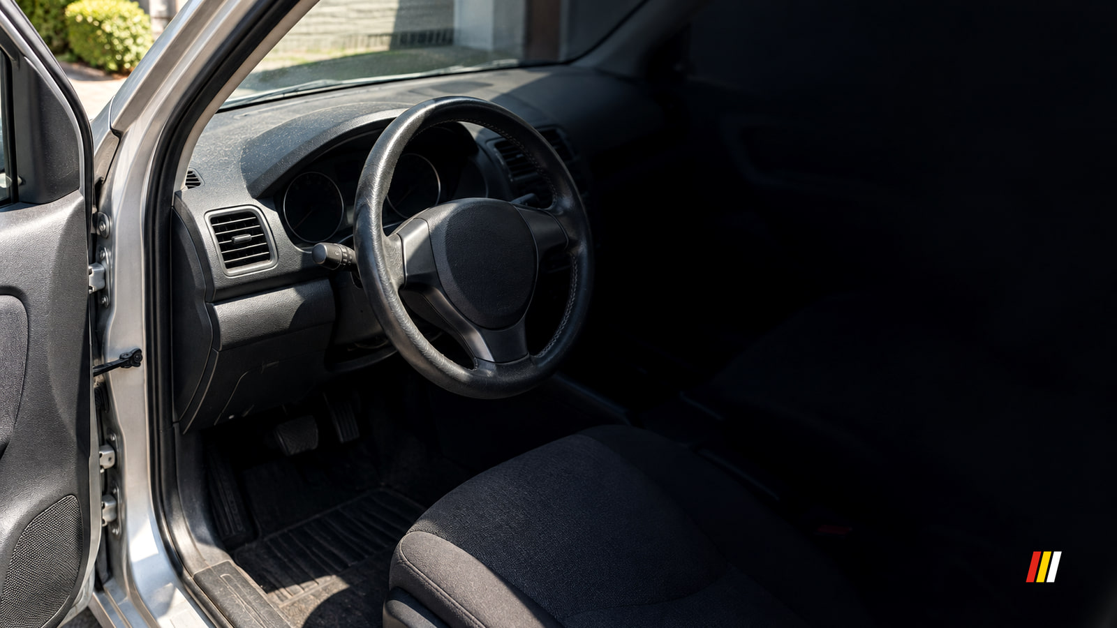 Steering wheel and a switched-off, dark instrument cluster seen through a used car's open driver's door, with hard midday sun on the door sill.