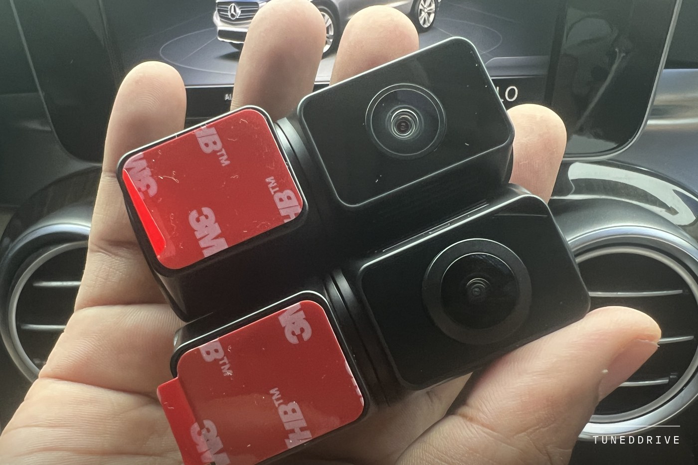 The A329S kit's two add-on cameras held in one hand, each with a red 3M adhesive pad on its mount, a car's centre screen behind