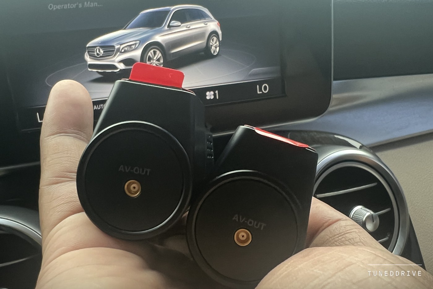 The backs of the two add-on cameras held in one hand, each showing a gold AV-OUT socket, with a car's centre screen and air vents behind