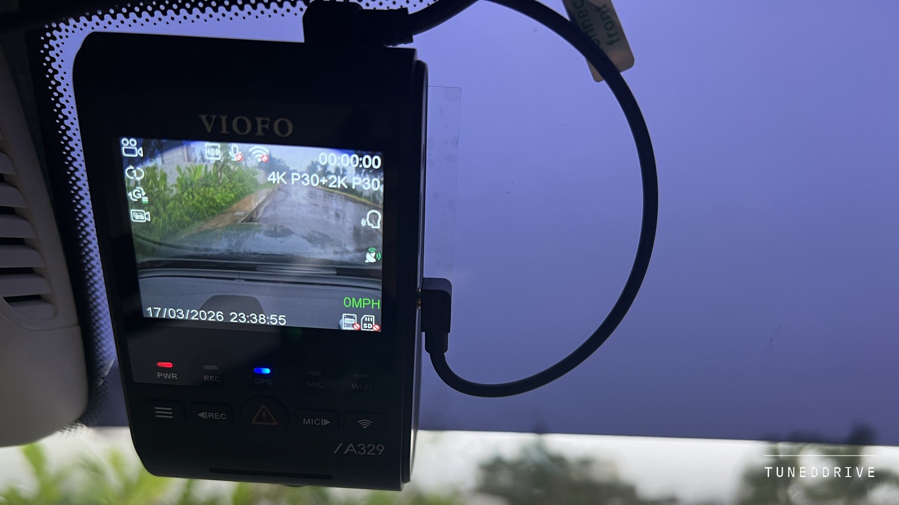 The VIOFO A329S front unit mounted high on a car windshield, its screen showing the live road view and recording status, the power cable looping to one side and a wet street through the glass