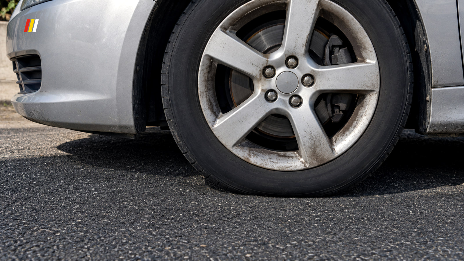 A silver car's front alloy wheel at a kerb in hard midday sun, with the hub, wheel nuts and brake disc visible through the open spokes.