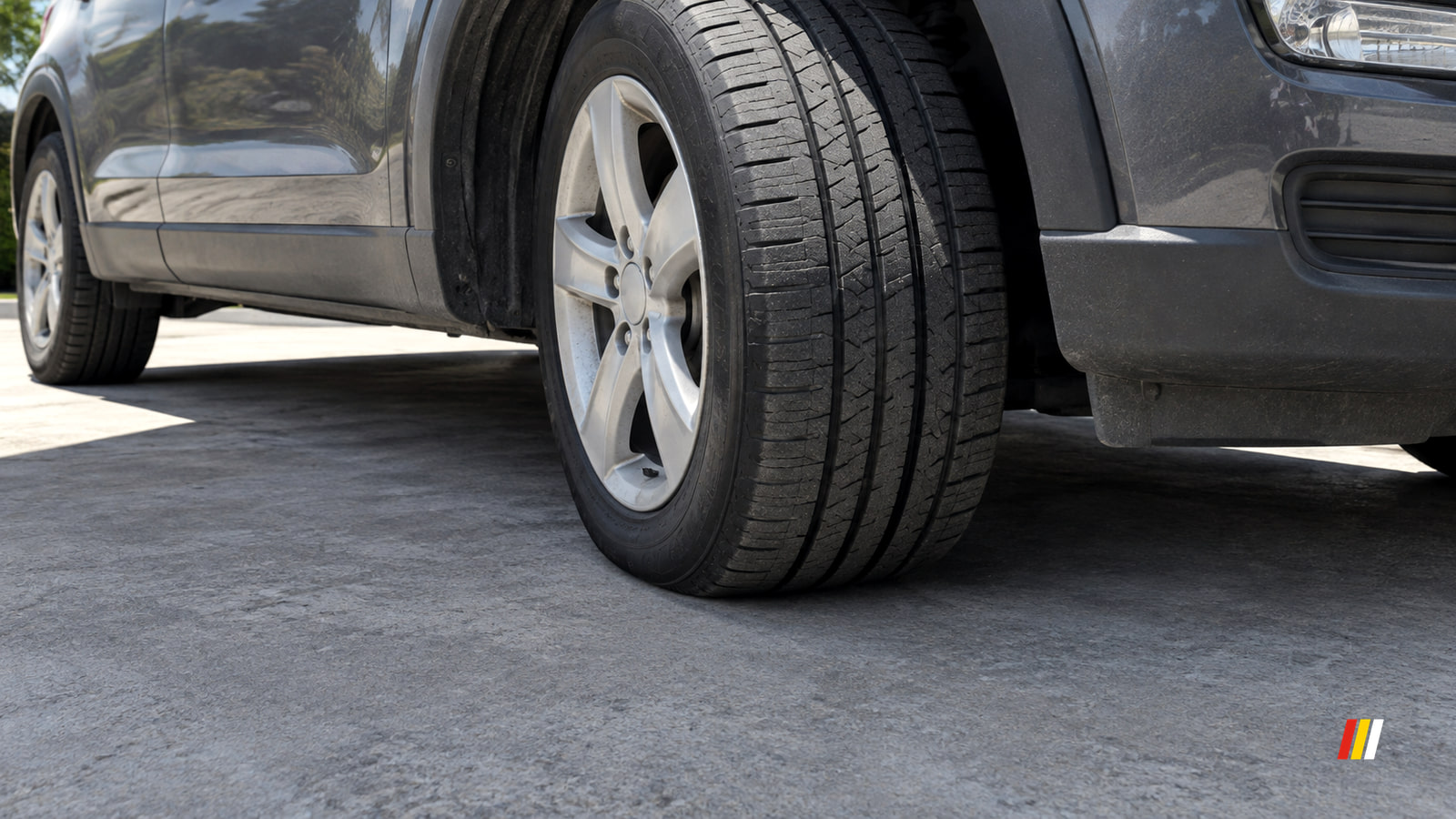 Turned front tyre of a dark grey SUV on a concrete driveway, its black sidewall and tread width seen low in hard midday sun.
