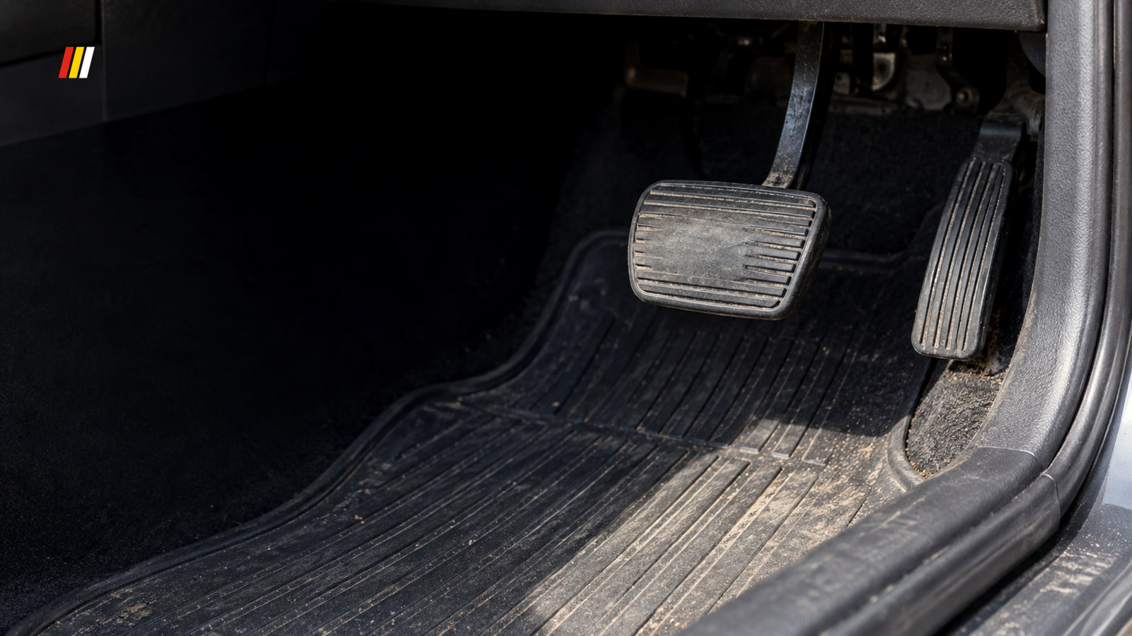 The worn black brake pedal and accelerator in a used car's footwell, seen low through the open driver's door in hard midday sun.