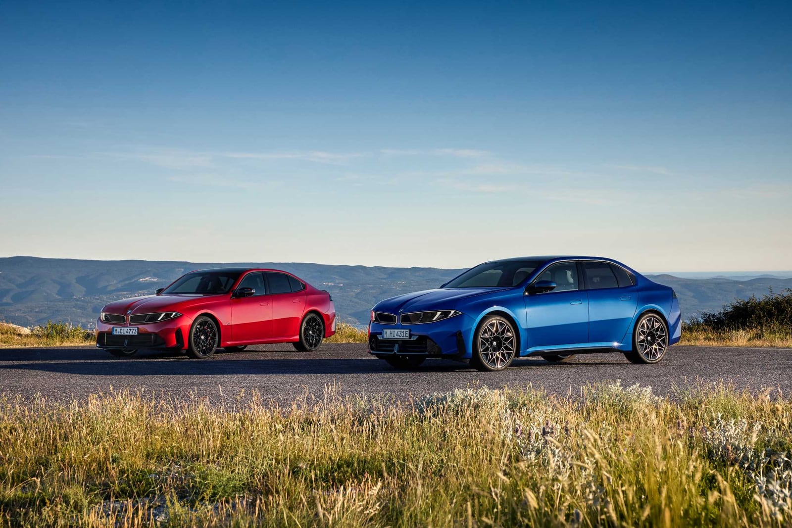 Red and blue BMW sedans parked on a paved road overlooking mountains under a clear blue sky.