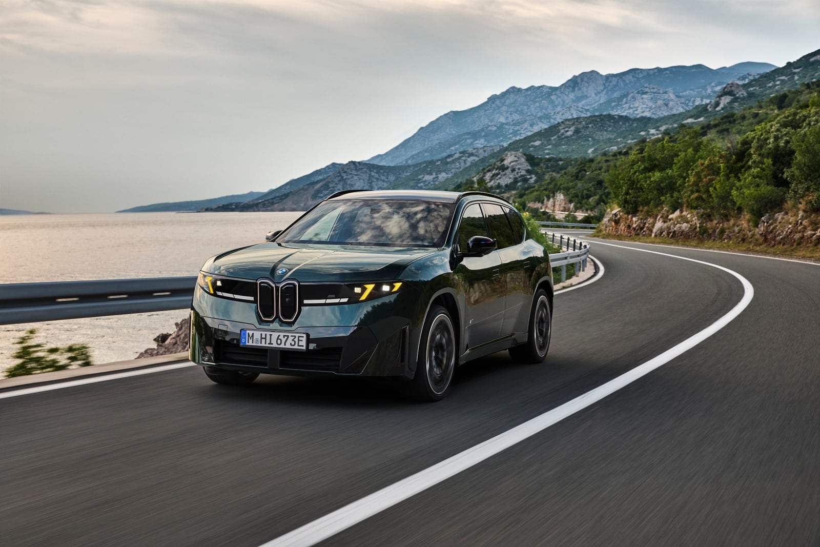 Dark green BMW SUV driving along a winding coastal road with mountains and water behind it.