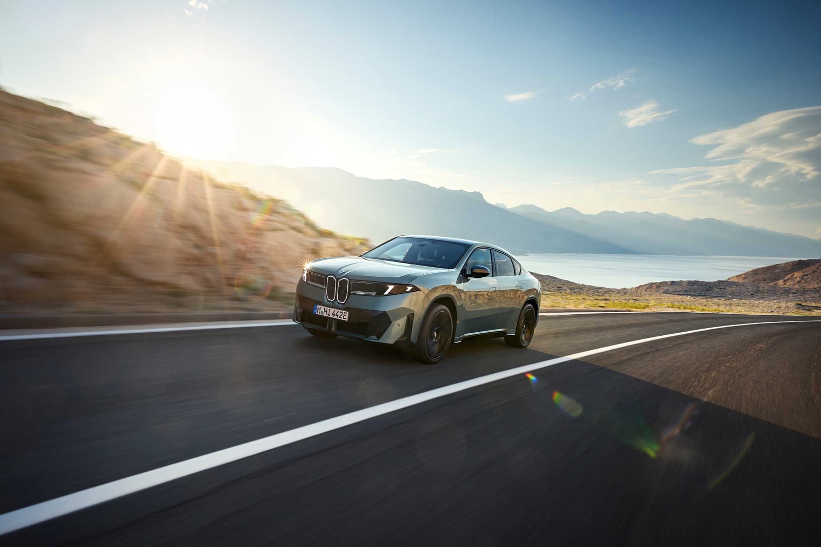 A silver BMW SUV coupe drives along a road beside water and mountains in sunlight.