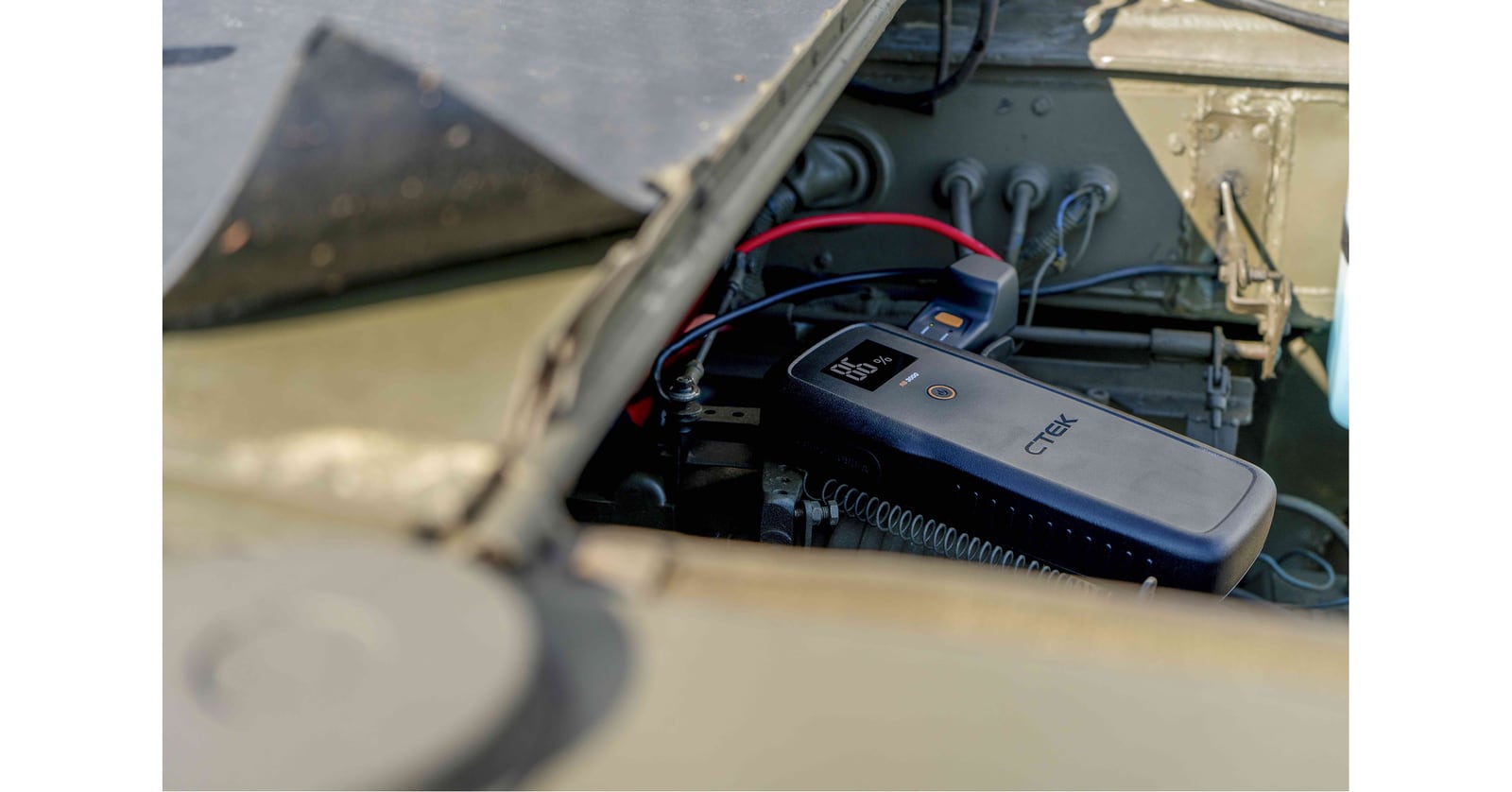 A CTEK portable booster rests in a vehicle engine bay with its display illuminated and cables attached.
