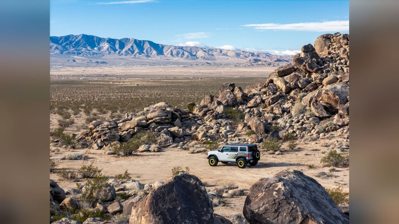 A silver Ford Bronco with lime wheel accents sits on a sandy trail among desert boulders.