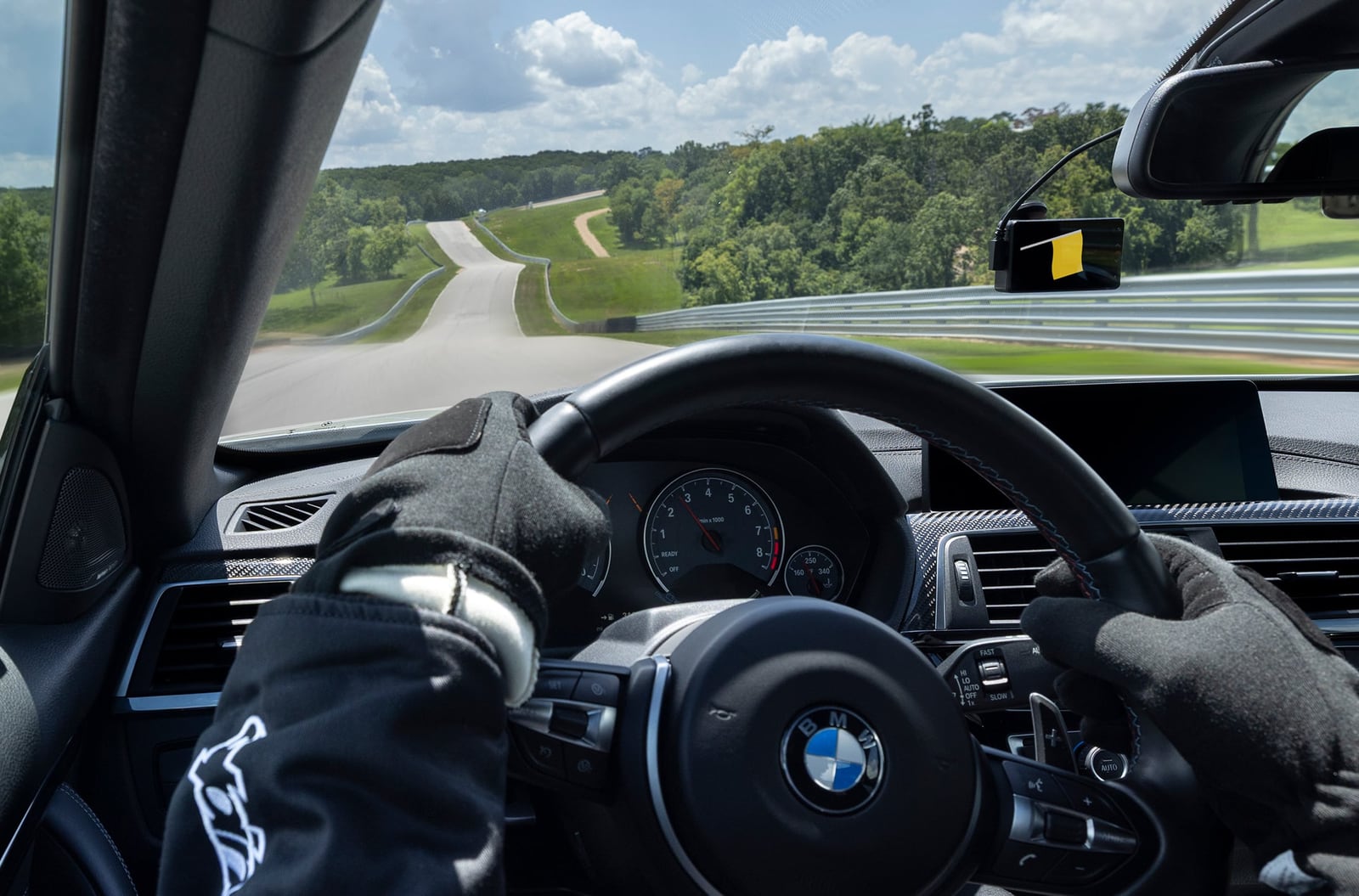 A windshield mounted screen shows a yellow flag above a BMW steering wheel as a driver navigates a racetrack.