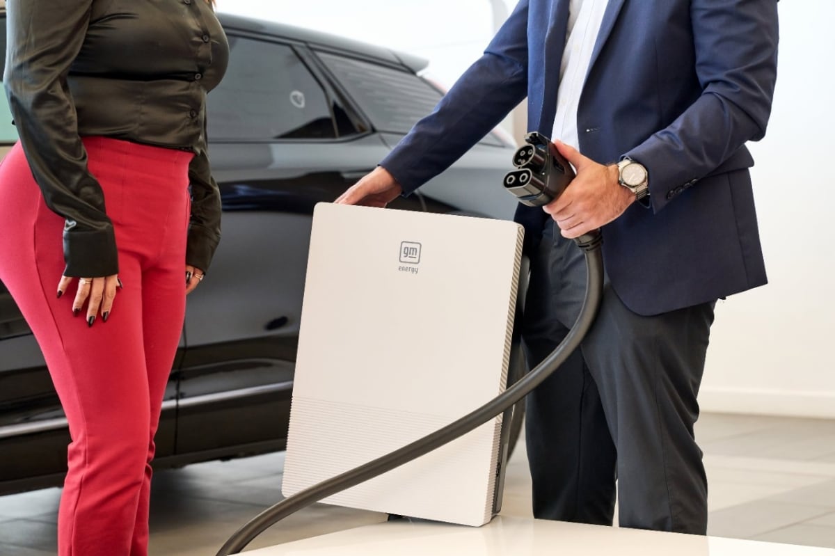 Two people display a GM Energy charging unit and connector beside a dark vehicle.