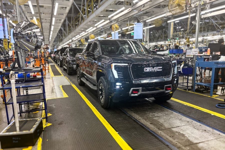 A dark GMC Sierra EV pickup faces forward on a factory assembly line with other trucks behind it.