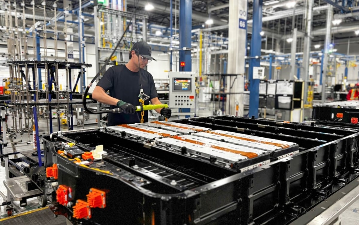 A worker holds a suspended assembly tool over silver modules in an open battery pack on a factory floor.