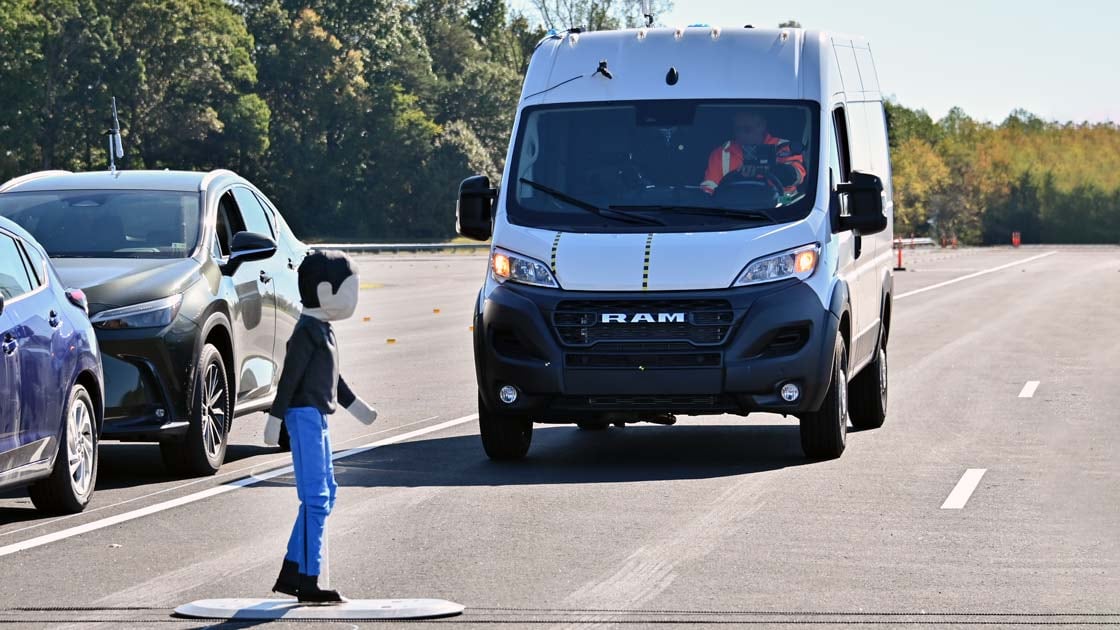 A white Ram cargo van approaches a pedestrian test dummy beside parked cars on a test track.