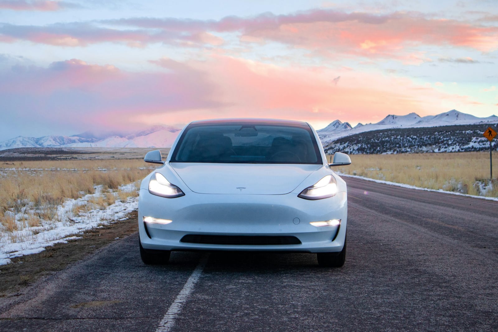 A white Tesla faces forward on a road beside snow covered mountains under a pink sky.