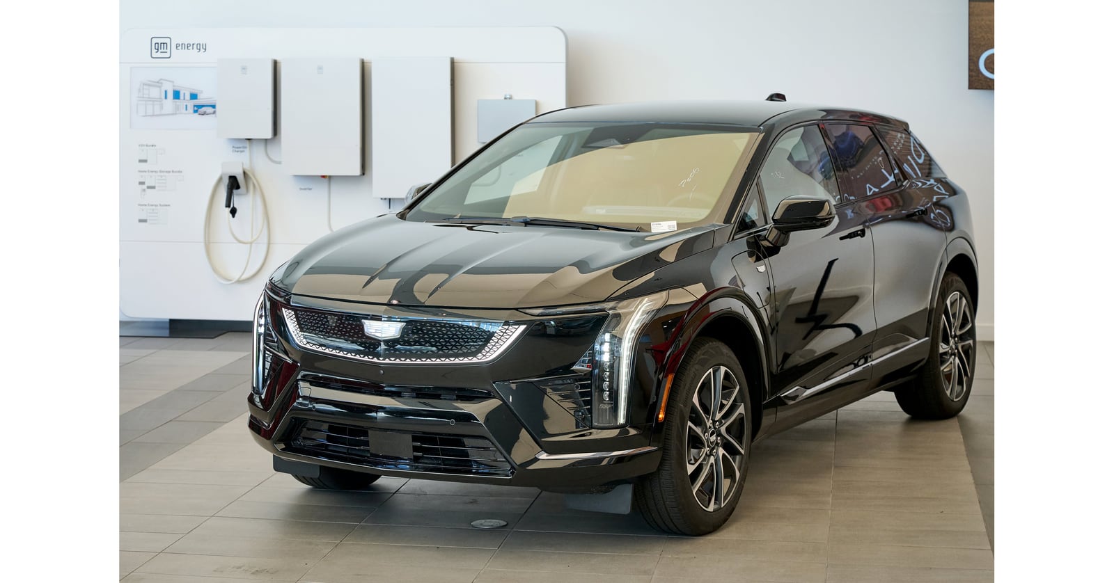 A black Cadillac SUV beside a GM Energy display with a charging cable and wall mounted home energy equipment.
