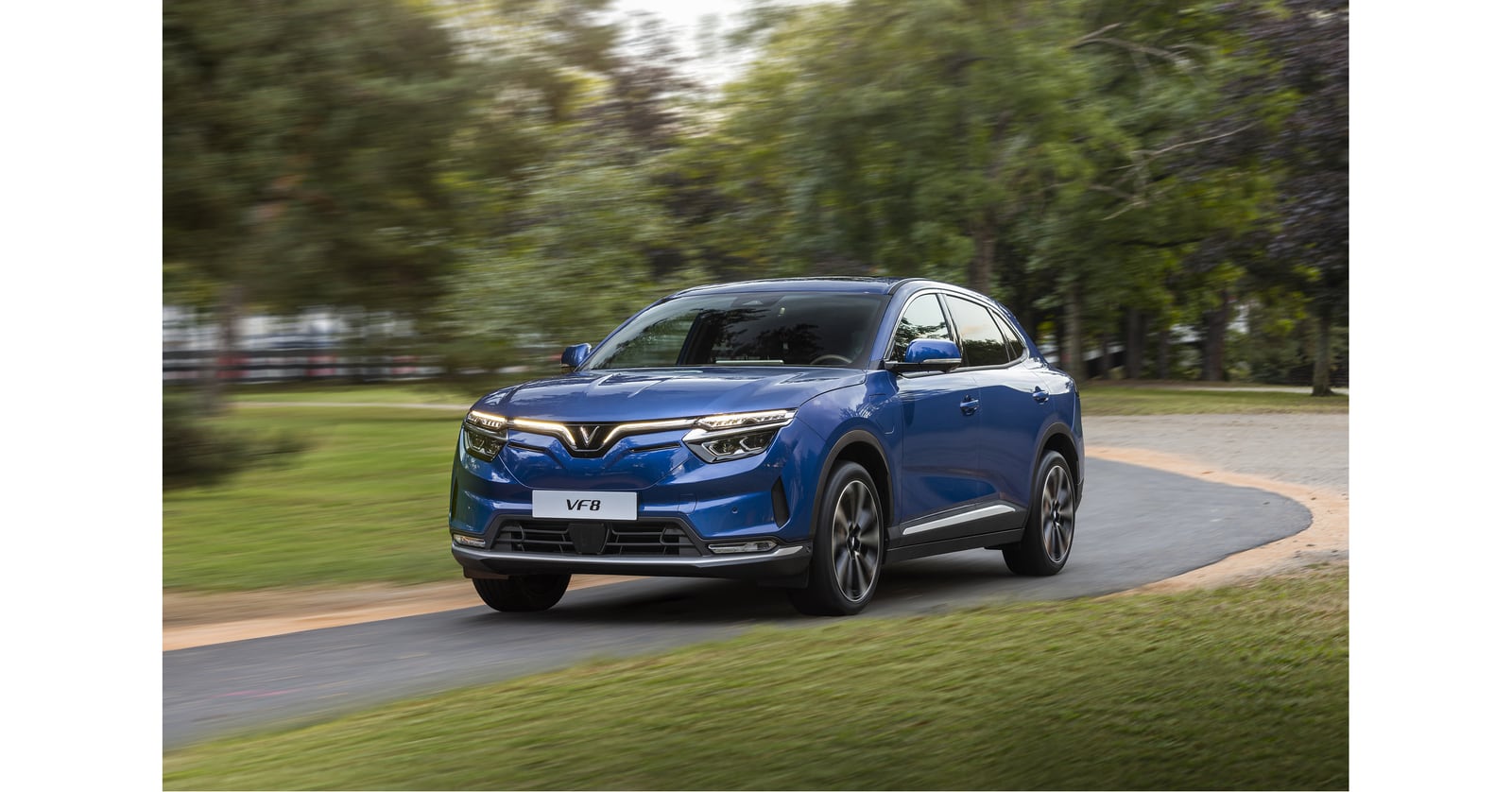 Blue VinFast VF 8 SUV driving along a curved road bordered by grass and trees.
