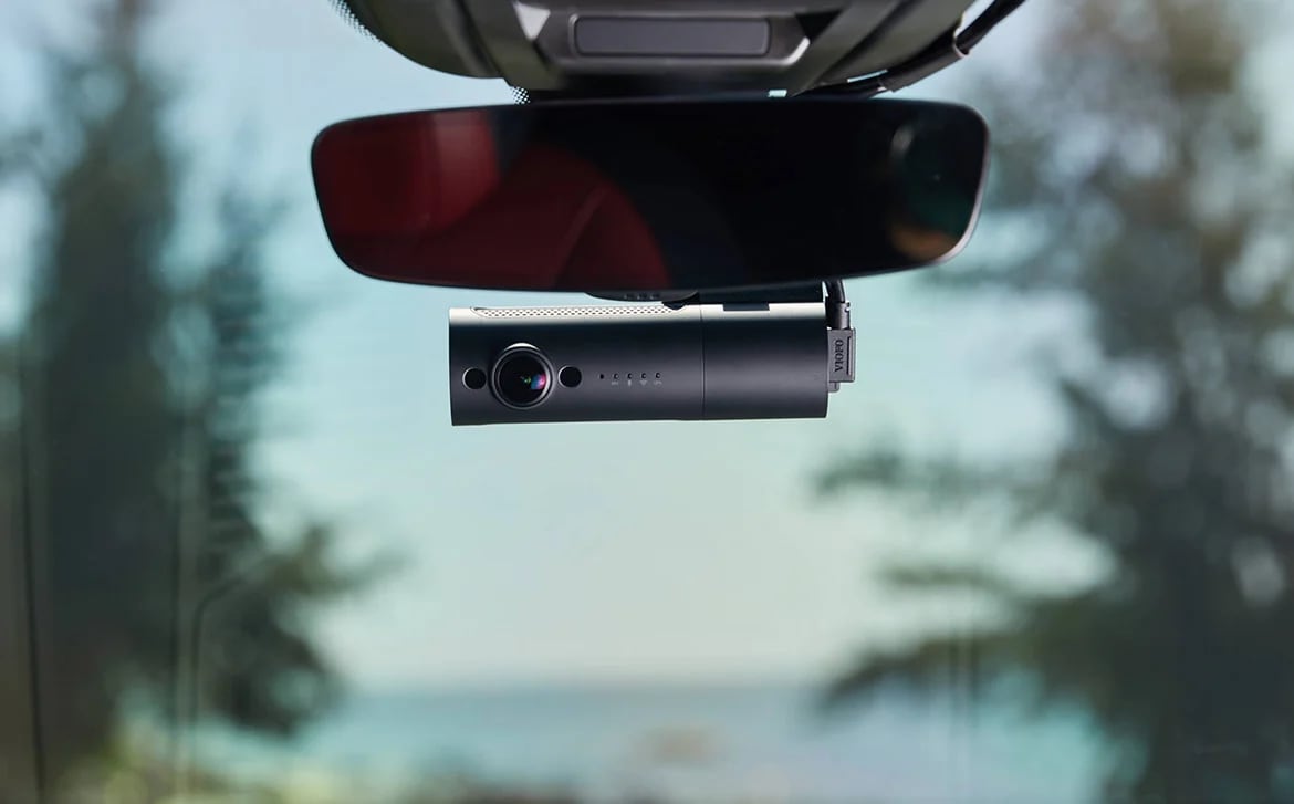 A VIOFO dashcam hangs beneath a car's rearview mirror, with blurred trees visible through the windshield.
